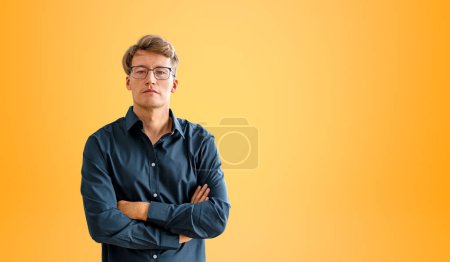 Téléchargez les photos : Homme d'affaires sérieux en chemise bleue et lunettes, regardant la caméra, bras croisés sur un fond orange vide. Concept de confiance, de leadership et de carrière - en image libre de droit