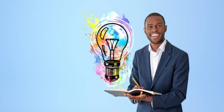 Photo for Portrait of cheerful young African American businessman in elegant suit holding notebook and pen and standing near blue wall with colorful lightbulb drawn on it. Concept of planning. Copy space - Royalty Free Image