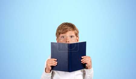 Téléchargez les photos : Portrait de petit garçon surpris couvrant son visage avec un livre debout sur fond bleu. Concept d'éducation, de lecture et d'acquisition de connaissances pour l'avenir - en image libre de droit