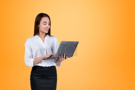 Photo for Smiling attractive businesswoman wearing formal wear is standing holding laptop near empty orange wall in background. Concept of model, successful business person, student, mobile gadget - Royalty Free Image