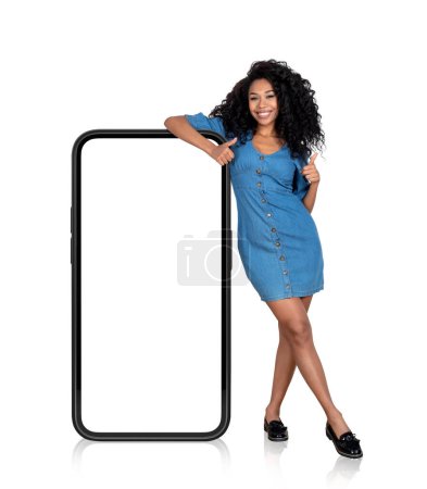 Téléchargez les photos : Femme noire souriante avec les pouces levés, debout près de grand smartphone avec écran d'espace de copie maquette, isolé sur fond blanc. Concept de médias sociaux - en image libre de droit