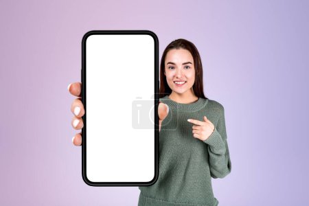 Téléchargez les photos : Femme souriante montrant et pointant du doigt vers un grand écran de smartphone d'espace de copie maquette, fond lilas. Concept de médias sociaux - en image libre de droit