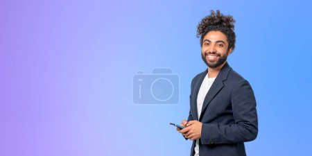Foto de Retrato de un joven y alegre empresario afroamericano en un elegante traje con un smartphone cerca de la pared púrpura. Concepto de comunicación empresarial. Copiar espacio - Imagen libre de derechos
