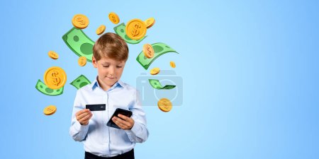 Photo for Adorable schoolboy holding smartphone and credit card standing near blue wall with dollar bills and coins around him. Concept of internet banking. Copy space - Royalty Free Image