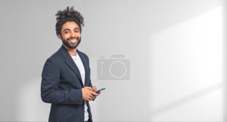 Foto de Retrato de un joven y alegre empresario afroamericano con un elegante traje que sostiene un smartphone de pie cerca de la pared blanca. Concepto de comunicación empresarial. Copiar espacio - Imagen libre de derechos
