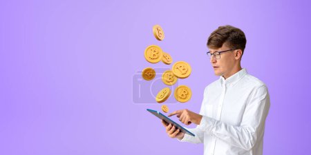 Téléchargez les photos : Jeune homme d'affaires européen utilisant un ordinateur tablette debout près du mur violet avec Bitcoins volant au-dessus. Concept de crypto-monnaie et d'applications bancaires sur Internet. Espace de copie - en image libre de droit