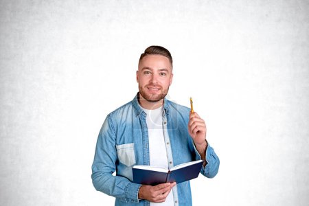 Téléchargez les photos : Portrait d'un étudiant souriant barbu dans des vêtements décontractés tenant un cahier et un stylo debout sur un fond de mur en béton. Concept d'éducation et de brainstorming - en image libre de droit