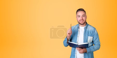 Foto de Retrato de estudiante universitario barbudo alegre en ropa casual sosteniendo cuaderno y pluma de pie sobre fondo amarillo. Concepto de educación y lluvia de ideas. Copiar espacio - Imagen libre de derechos