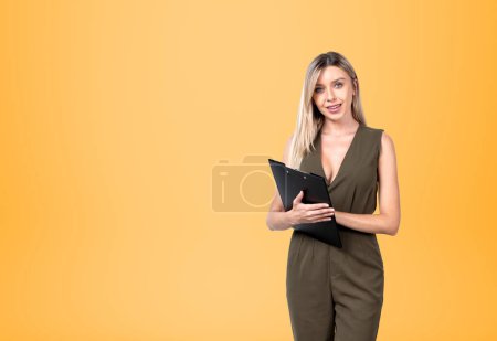 Photo for Smiling attractive businesswoman wearing formal wear is standing holding clipboard near empty orange wall in background. Concept of model, business person, student, secretary, time management - Royalty Free Image