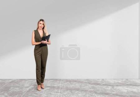 Photo for Smiling attractive businesswoman wearing formal wear is standing holding clipboard near empty white wall in background. Concrete tile floor. Concept of model, business person, student, secretary - Royalty Free Image