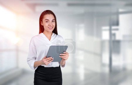 Photo for Smiling attractive businesswoman wearing formal wear is standing holding tablet device at office workplace in background. Concept of model, successful business person, employee, mobile gadget - Royalty Free Image