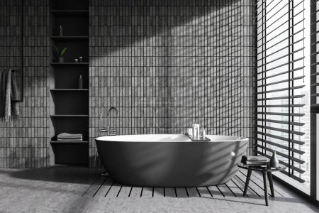 Téléchargez les photos : Intérieur de salle de bain élégante avec des murs de tuiles grises, sol en béton, confortable baignoire grise debout près de la fenêtre panoramique avec stores et étagères avec des produits d'hygiène. Rendu 3d - en image libre de droit