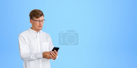 Téléchargez les photos : Portrait du jeune homme d'affaires européen serioius en chemise blanche regardant l'écran du smartphone debout sur fond bleu. Concept de médias sociaux et de communication. Espace de copie - en image libre de droit
