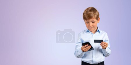 Téléchargez les photos : Portrait d'adorable petit garçon en vêtements formels tenant smartphone et carte de crédit debout sur fond violet. Concept de banque en ligne et d'achats en ligne. Espace de copie - en image libre de droit