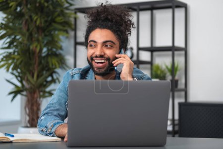 Foto de Feliz hombre de negocios del oeste oriental llamando por teléfono, portátil y portátil en el escritorio. Concepto de conexión y comunicación - Imagen libre de derechos