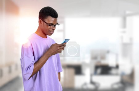 Photo for African American businessman wearing casual wear is standing holding smartphone at office workplace in background. Concept of working process, texting message for colleagues, student, mobile gadget - Royalty Free Image