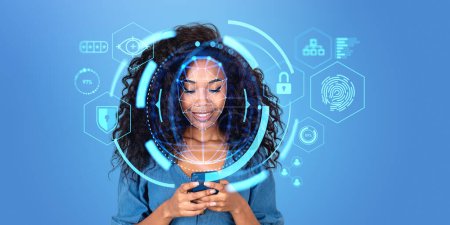 Téléchargez les photos : Joyeux jeune femme d'affaires afro-américaine utilisant un smartphone avec interface immersive de reconnaissance faciale debout sur fond bleu. Concept d'authentification biométrique - en image libre de droit