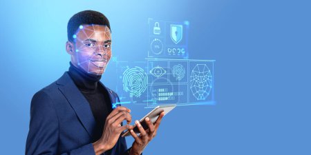 Téléchargez les photos : Portrait d'un jeune homme d'affaires afro-américain souriant utilisant une tablette avec une interface de balayage biométrique immersive sur fond bleu. Espace de copie - en image libre de droit
