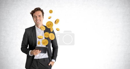 Photo for Smiling young European businessman holding tablet with dollar coins floating above it standing near concrete wall. Concept of internet banking and online transactions. Copy space - Royalty Free Image