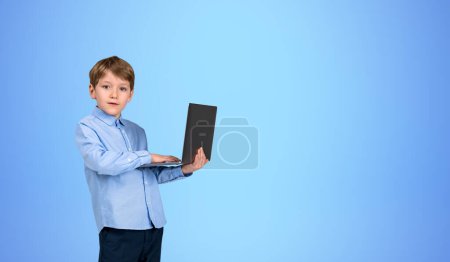 Foto de Retrato de niño concentrado en ropa formal sosteniendo el portátil de pie sobre fondo azul. Concepto de e-learning y redes sociales. Tecnología moderna y conexión a Internet. Copiar espacio - Imagen libre de derechos