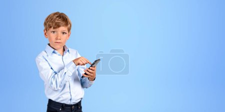 Téléchargez les photos : School boy doigt toucher le téléphone, portrait sérieux en regardant l'appareil photo sur fond bleu vide espace de copie. Concept d'application mobile, d'éducation en ligne et de shopping - en image libre de droit