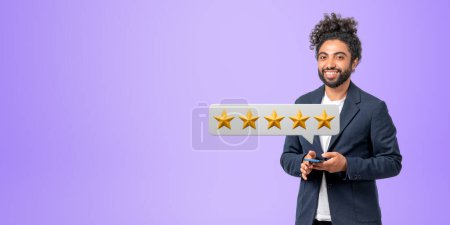 Smiling Middle Eastern businessman in suit holding smartphone and giving five star rating. Purple background. Concept of service and product feedback and ranking. Copy space