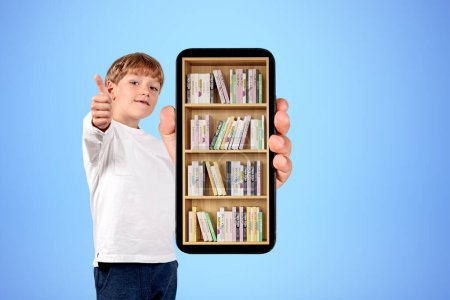 Téléchargez les photos : Enfant garçon montrant pouce levé et grand écran de téléphone avec bibliothèque numérique sur fond bleu. Concept d'éducation et d'apprentissage en ligne - en image libre de droit