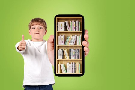 Photo for Handsome boy in casual wear is showing smartphone case with digital library with approval. Bookshelf with various books. Concept of e-learning and online education. Green wall in background - Royalty Free Image