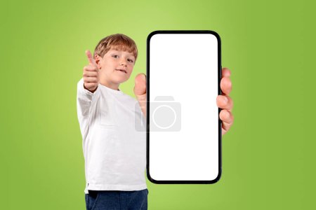 Photo for Kid boy smiling, showing thumb up and smartphone with large mock up copy space screen on green background. Concept of e-learning - Royalty Free Image