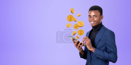 Foto de Sonriente joven empresario afroamericano sosteniendo la tableta con monedas de dólar flotando sobre ella de pie cerca de la pared púrpura. Concepto de banca por Internet y transacciones en línea. Copiar espacio - Imagen libre de derechos