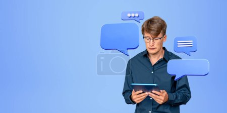 Photo for Portrait of concentrated young European businessman holding tablet with speech bubbles and social media notifications around him. Online communication and chatting concept. Blue background, copy space - Royalty Free Image