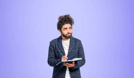 Photo for Thoughtful young Middle Eastern man college student standing with notebook and pen and looking sideways and upwards over purple background. Concept of planning, creativity and education - Royalty Free Image