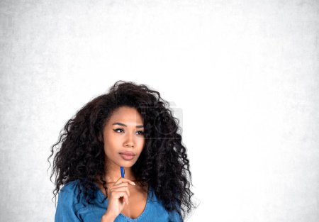 Téléchargez les photos : Une femme d'affaires afro-américaine séduisante et réfléchie portant des vêtements formels se tient debout en touchant le menton avec un stylo près d'un mur blanc en béton vide en arrière-plan. Concept de modèle, homme d'affaires prospère - en image libre de droit