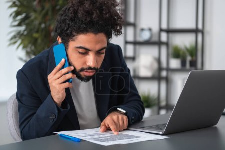 Photo for Arab businessman calling on the phone, look through the paper contract on desk with laptop. Concept of career and recruitment. - Royalty Free Image