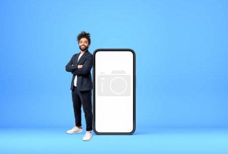 Photo for West eastern businessman smiling, standing full length near large smartphone mock up empty screen, blue background. Concept of online network and social media - Royalty Free Image