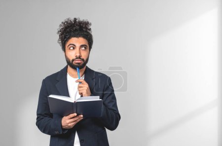 Photo for Pondering handsome businessman wearing formal wear is standing touching his chin with pen holding notebook near empty white wall in background. Concept of model, thinking for creative idea - Royalty Free Image