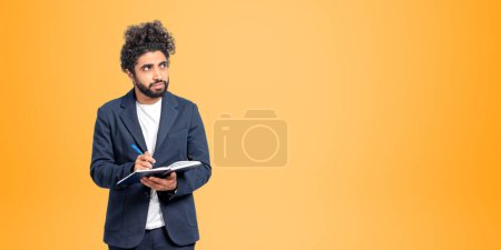 Photo for Pensive young Middle Eastern man college student standing with notebook and pen and looking sideways and upwards over orange background. Concept of planning, creativity and education. Copy space - Royalty Free Image