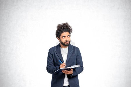 Photo for Pensive young Middle Eastern man college student standing with notebook and pen and looking sideways and upwards over concrete background. Concept of planning, creativity and education - Royalty Free Image
