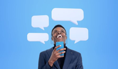 Téléchargez les photos : Homme d'affaires africain riant avec téléphone à la main, bulles de message texte maquette sur fond bleu. Concept de médias sociaux, conversation d'affaires et réseau - en image libre de droit