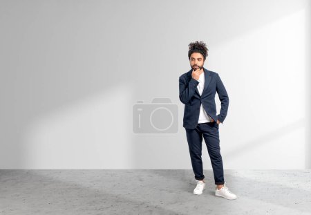 Photo for Middle eastern businessman full length standing with thoughtful look, hand to chin. Office room with grey concrete floor. Copy space blank wall background. Concept of startup - Royalty Free Image