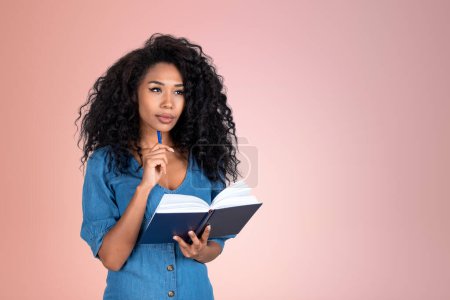 Photo for Thoughtful attractive African American businesswoman wearing formal wear is standing touching chin with pen hoding notebook near empty pale pink wall in background. Concept of model, learning student - Royalty Free Image