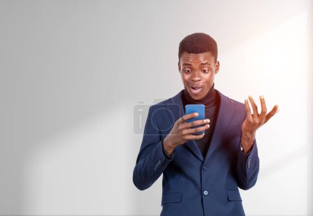 Téléchargez les photos : Beau homme d'affaires afro-américain en costume formel est debout en regardant le smartphone près du mur blanc vide en arrière-plan. Concept de gadgets modernes, communication mobile, gestion du temps - en image libre de droit