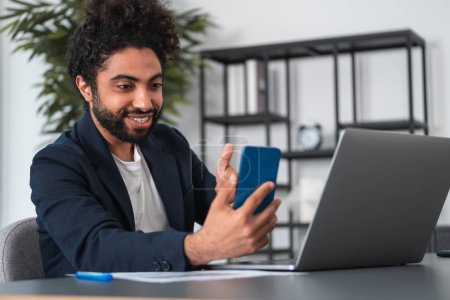 Foto de Hombre de negocios árabe sonriente hablando en una videoconferencia de teléfono móvil, contrato y portátil en el escritorio. Mostrando el pulgar hacia arriba. Concepto de éxito y carrera - Imagen libre de derechos
