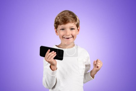 Téléchargez les photos : Happy enfant garçon portrait avec un poing levé et dispositif à la main, copier l'espace fond violet vide. Concept d'études et de réalisations - en image libre de droit