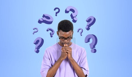 Téléchargez les photos : Portrait d'un jeune homme africain réfléchi étudiant en vêtements décontractés debout près du mur bleu avec des points d'interrogation et de réfléchir dur. Concept de choix de carrière et de prise de décision - en image libre de droit