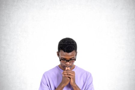 Téléchargez les photos : Portrait d'un jeune homme africain étudiant en vêtements décontractés et lunettes réfléchissant debout sur un fond de béton. Concept de résolution de problèmes et choix de carrière - en image libre de droit