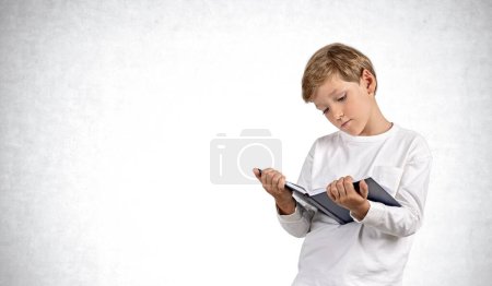 Photo for Smart boy portrait with book in hands reading, copy space grey concrete wall background. Concept of online education, knowledge, literature and studies - Royalty Free Image