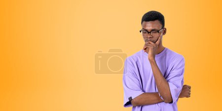 Foto de Africano pensativo estudiante hombre en gafas y camisa púrpura, reflexivo mirando hacia abajo en el espacio de copia fondo naranja vacío. Concepto de dudas, ideas e inseguridad - Imagen libre de derechos