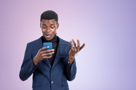 Photo for Handsome African American businessman in formal suit is standing watching at smartphone near empty gradient purple wall in background. Concept of modern gadgets, mobile communication, time management - Royalty Free Image