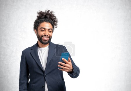 Foto de Retrato de hombre de negocios de Oriente Medio con teléfono inteligente en la mano, comunicación en línea y red sobre fondo de pared de hormigón gris. Concepto de internet global. Copiar espacio - Imagen libre de derechos